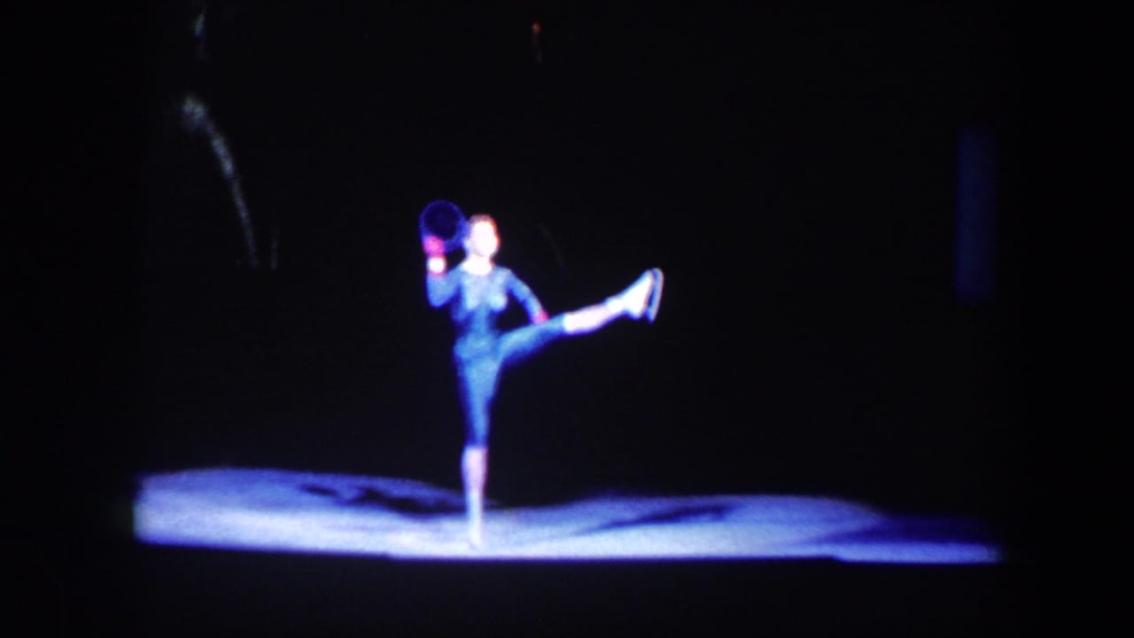 1960 Baltimore 8mm frame showing a solo stage dancer in a dark costume lifting one leg in a bright pool of light.