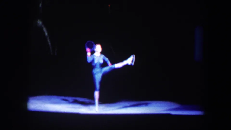 1960 Baltimore 8mm frame showing a solo stage dancer in a dark costume lifting one leg in a bright pool of light.