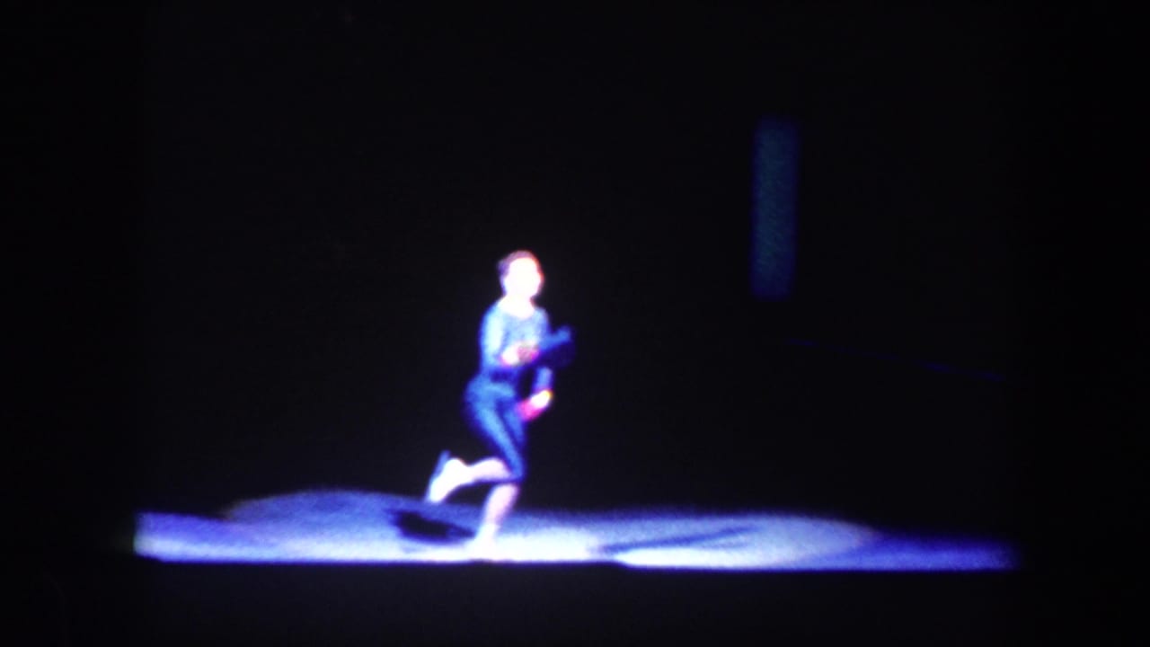 Solo performer kneels or poses in a spotlight on a dark stage.