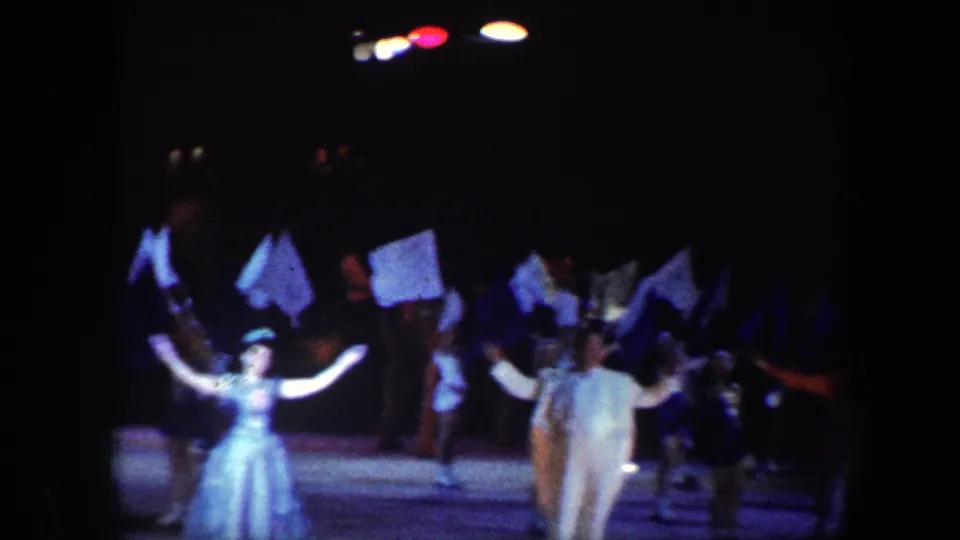 1960 Baltimore 8mm frame showing performers in costumes raising flags on a dark stage under colored spotlights.