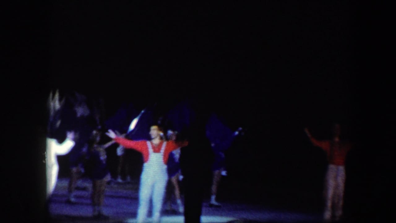 A performer in red and white stands with arms out as others move behind flags.