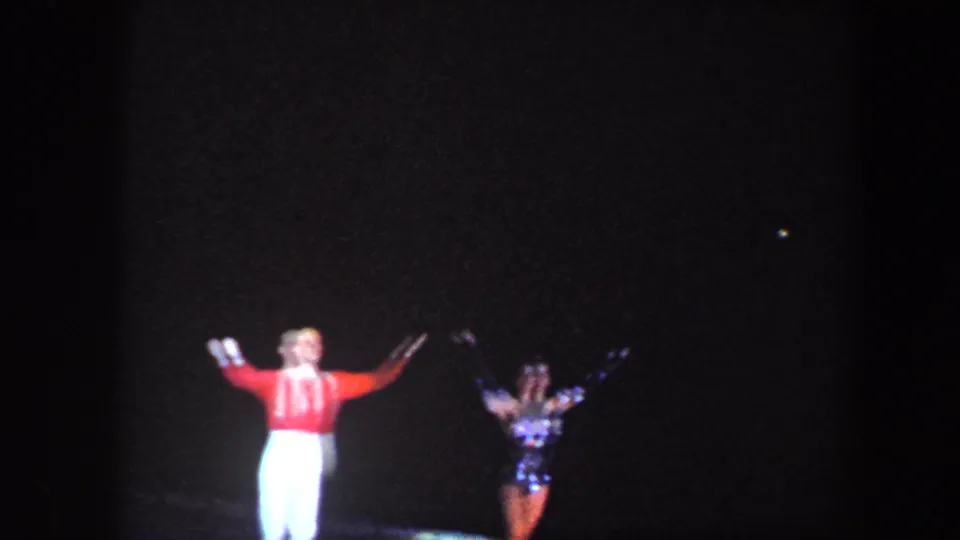 Low-light 1960 Baltimore 8mm frame with costumed performers, including a red-and-white outfit, on a dark stage.