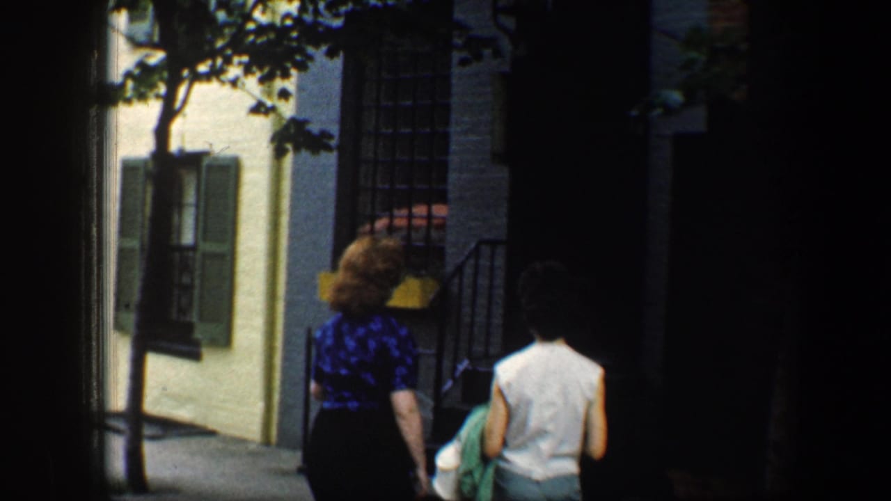 Two women walk away beside row houses and trees.