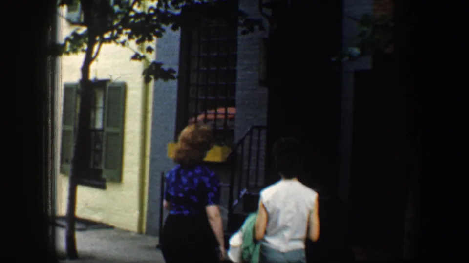 Two adult women walk away on a Baltimore sidewalk beside shuttered row houses and leafy trees, circa 1960 per clip metadata.