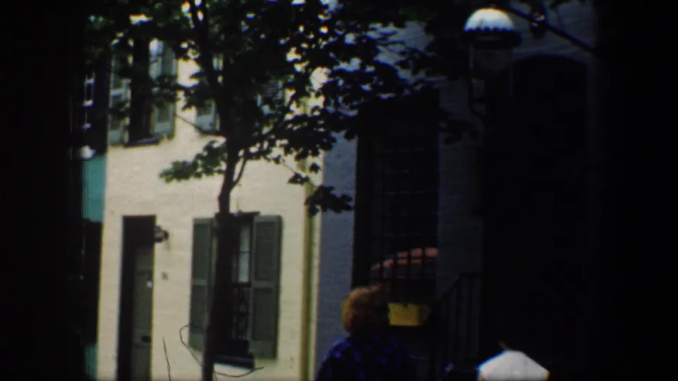 Two adult women continue walking away through a shaded Baltimore residential block with shutters, windows, and tree cover visible.
