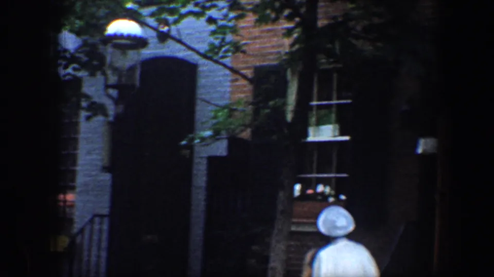 1960 Baltimore home-movie frame with brick row houses, shaded stoops, windows, and a small pedestrian wearing a white hat.