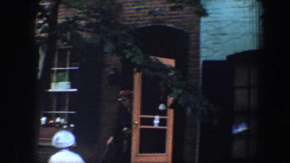 1960 Baltimore frame showing brick and teal row-house fronts, open doors, shaded windows, and pedestrians along the sidewalk.