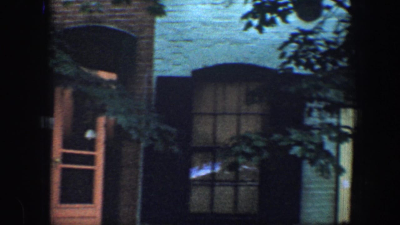 1960 Baltimore home-movie frame showing an orange entrance door, dark shutters, windows, and leafy branches across a residential exterior.