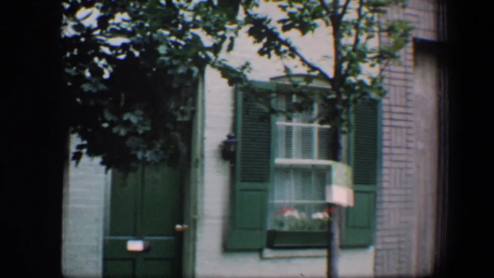 1960 Baltimore home-movie frame with green shutters, a window box with flowers, a green door, brickwork, and overhanging leaves.