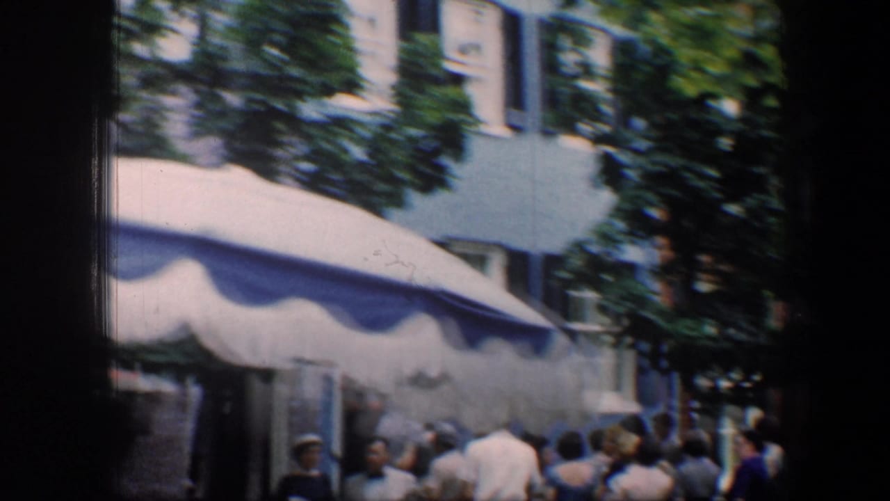 1960 Baltimore 8mm frame showing people gathered near a blue-and-white awning, trees, and pale building facades.