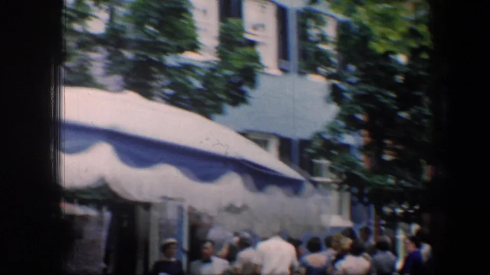1960 Baltimore 8mm frame showing people gathered near a blue-and-white awning, trees, and pale building facades.