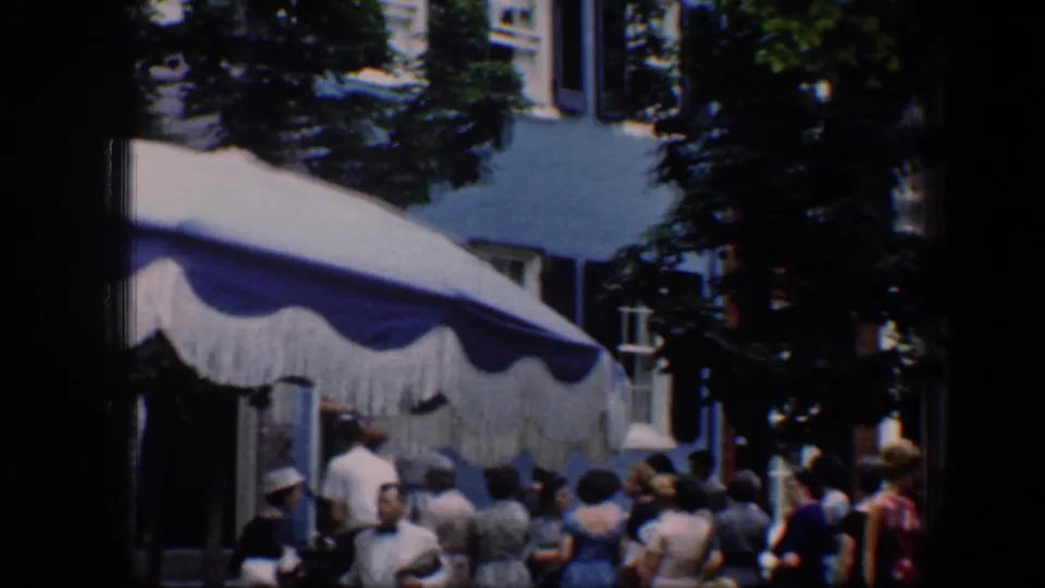 1960 Baltimore street view with a sidewalk crowd beneath a scalloped awning, framed by trees and upper-story windows.
