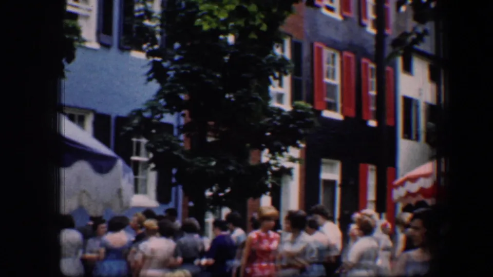 1960 Baltimore home-movie frame showing people gathered near shuttered buildings and a red-and-white striped umbrella.