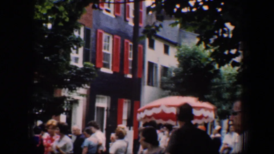 1960 Baltimore street scene with people standing near red-shuttered facades, trees, and a red-and-white striped umbrella.