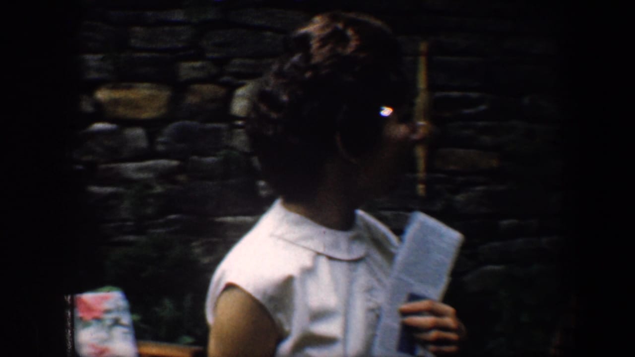 Woman in a white sleeveless dress holding a booklet near a stone wall.