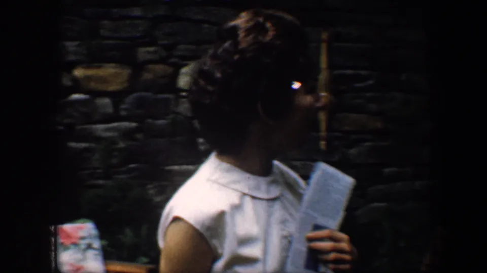 A woman in a white sleeveless dress holds a folded booklet or paper near a stone wall in 1960 Baltimore home-movie footage.