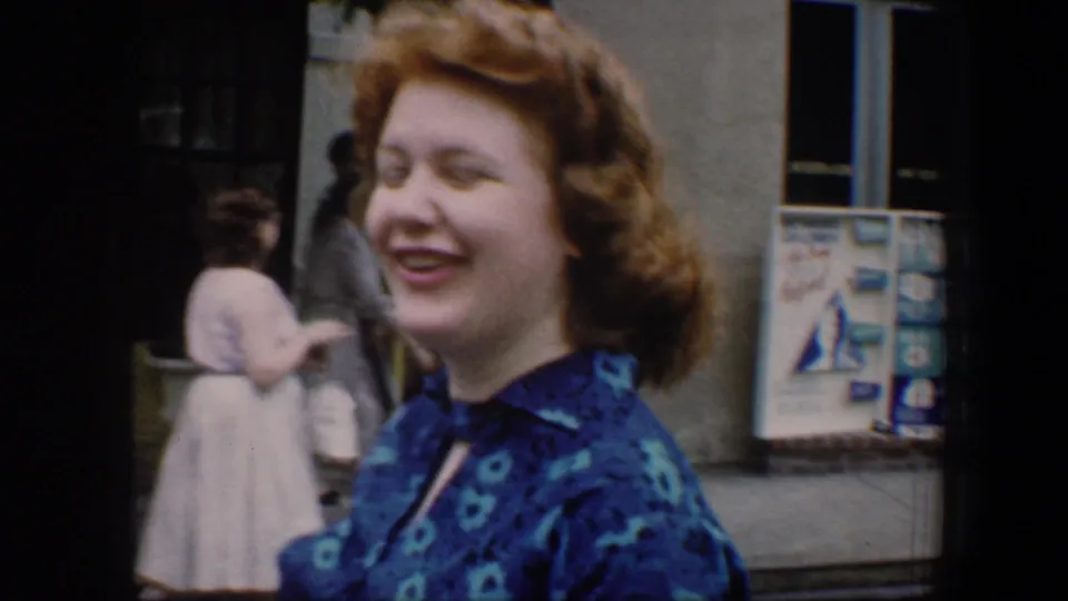A smiling woman in a blue floral dress stands on a sidewalk, with other pedestrians and a postered wall behind her.