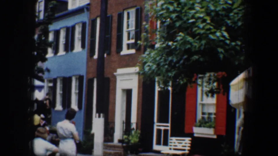 Painted Baltimore row houses with black shutters and a leafy street tree appear with people gathered near the sidewalk.