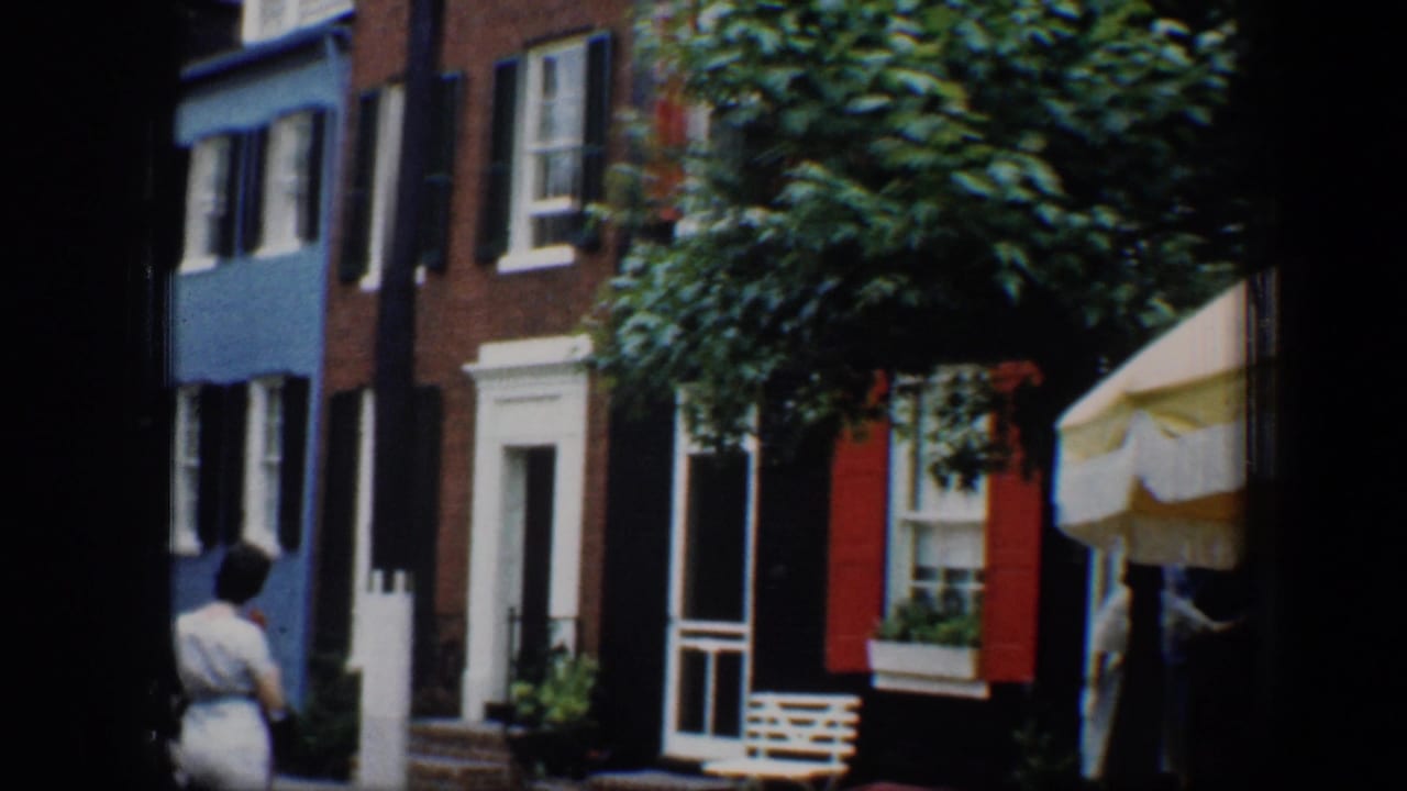 1960 Baltimore street view with colorful row houses, shuttered windows, a red door, and a pedestrian at the left edge.
