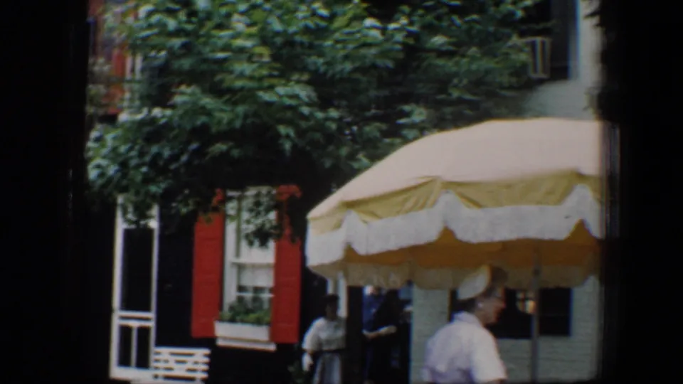 1960 Baltimore street frame showing a yellow-and-white scalloped umbrella, shuttered row houses, and adults near the sidewalk.