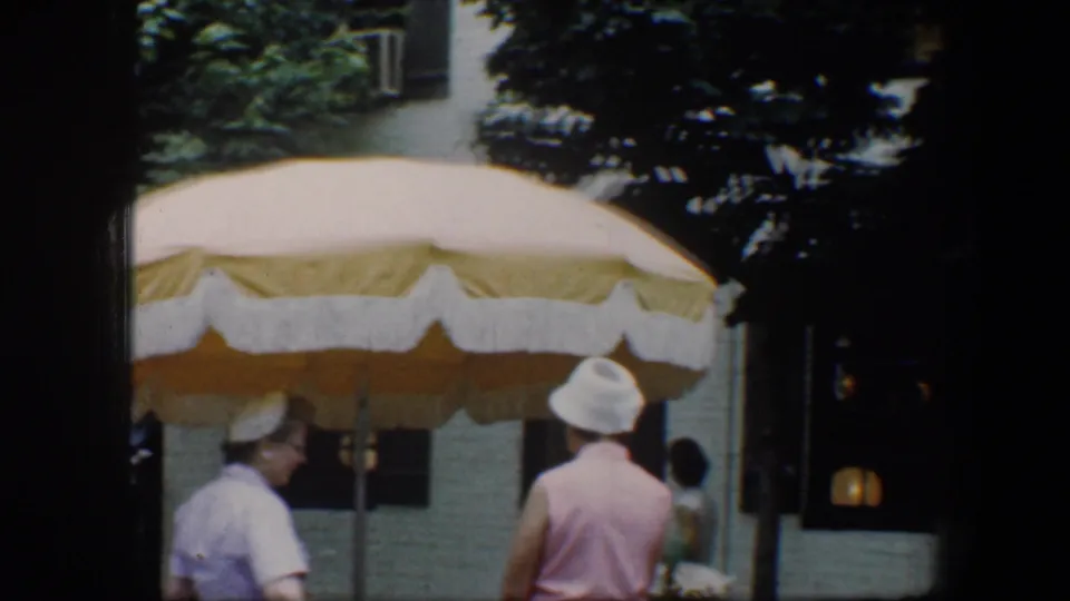 1960 Baltimore home-movie frame with two adults near a large yellow-and-white umbrella and pastel row-house facades.