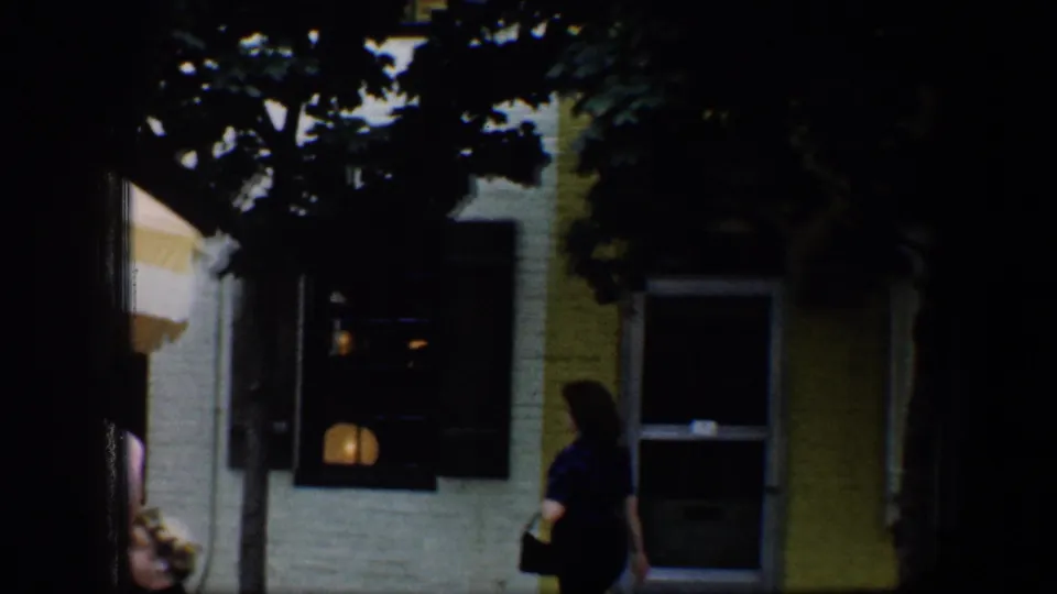 1960 Baltimore street view with a pedestrian, shaded row-house windows, dark shutters, and part of a yellow umbrella at the left.