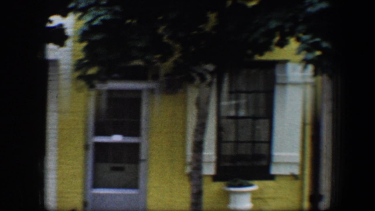 1960 Baltimore 8mm frame showing a yellow house front with white trim, dark window shutters, doorway, and tree shadow across the facade.