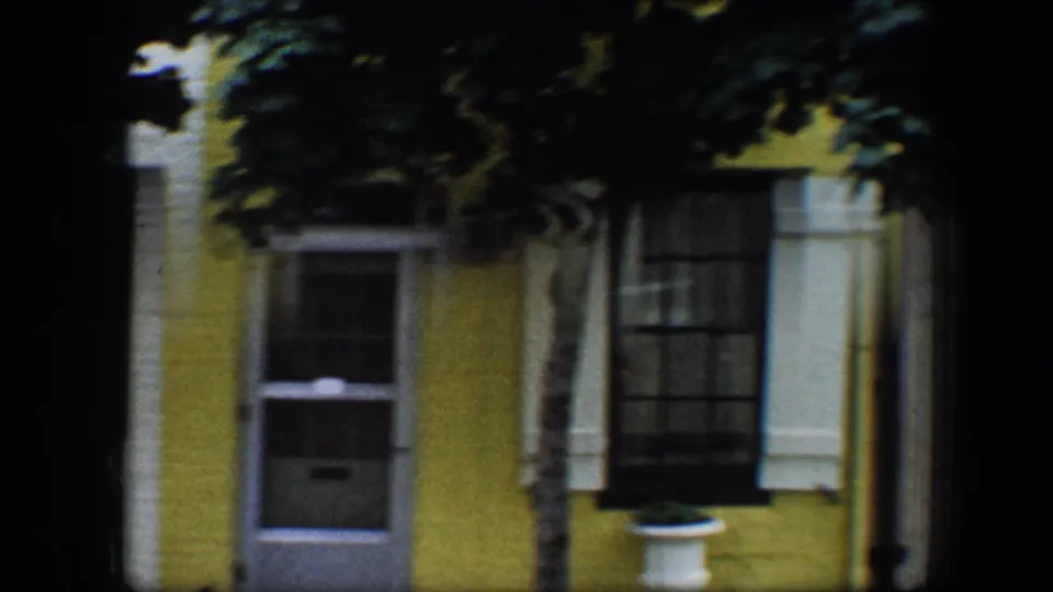 1960 Baltimore 8mm frame showing a yellow house front with white trim, dark window shutters, doorway, and tree shadow across the facade.