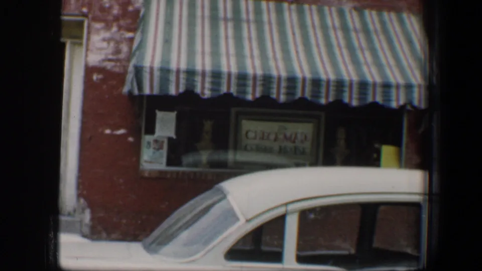 1960 Baltimore street view with a striped storefront awning, partially readable shop sign, brick wall, and classic white car parked in front.