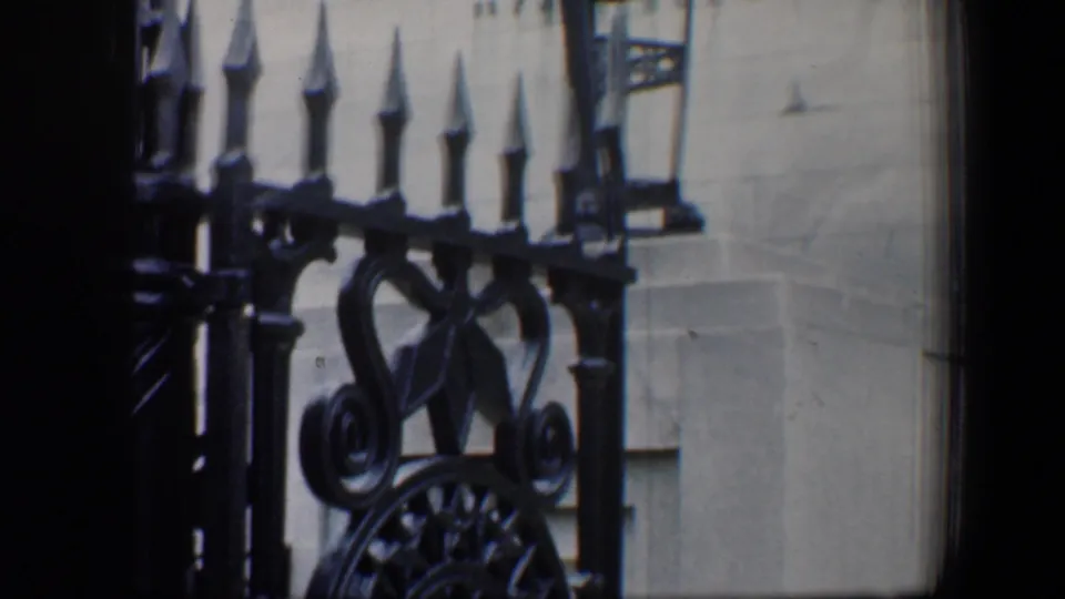 Close 8mm view of ornate black wrought iron fencing against a light stone wall, with architectural details partly obscured by soft focus.
