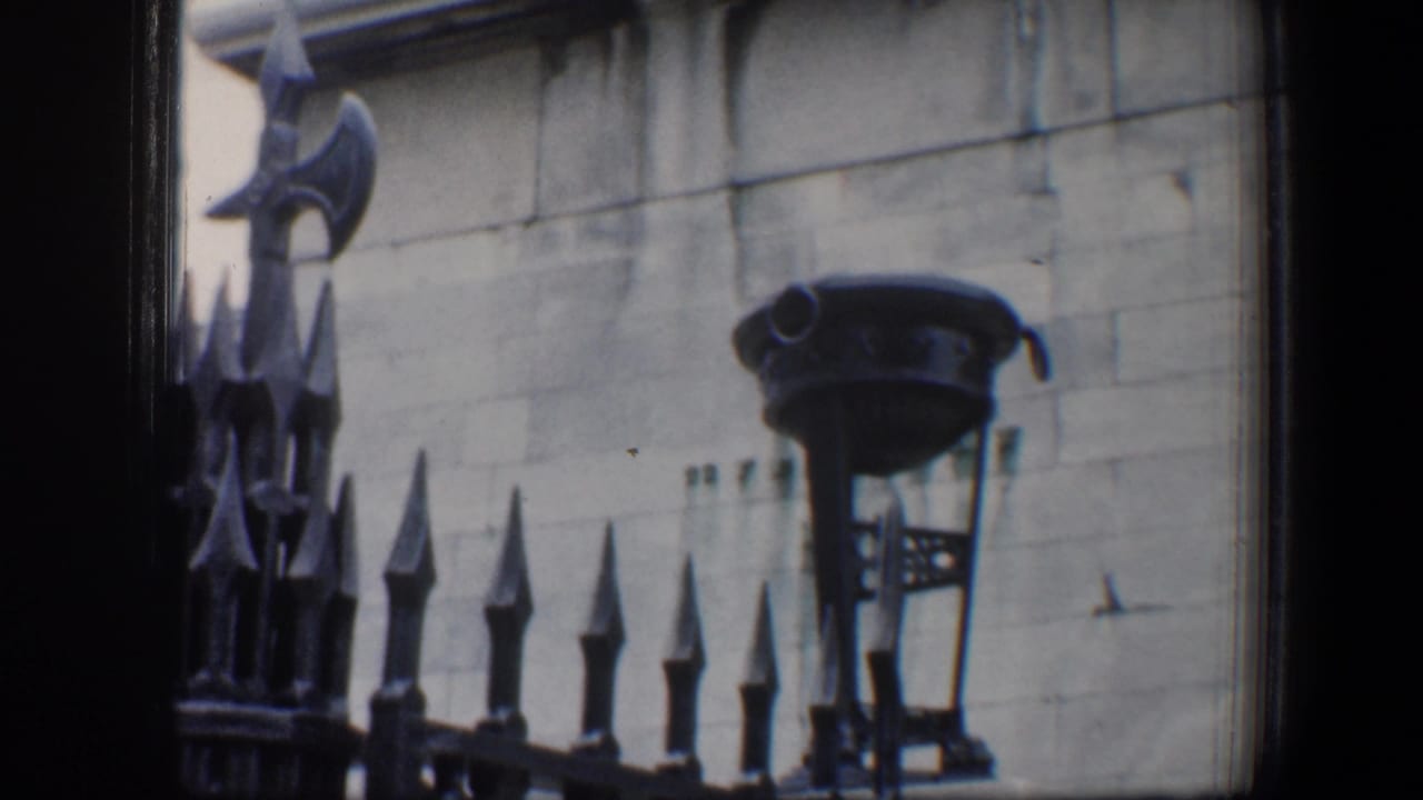 1960 Baltimore home-movie frame showing black iron fencing and a decorative lamp mounted near a pale stone facade.