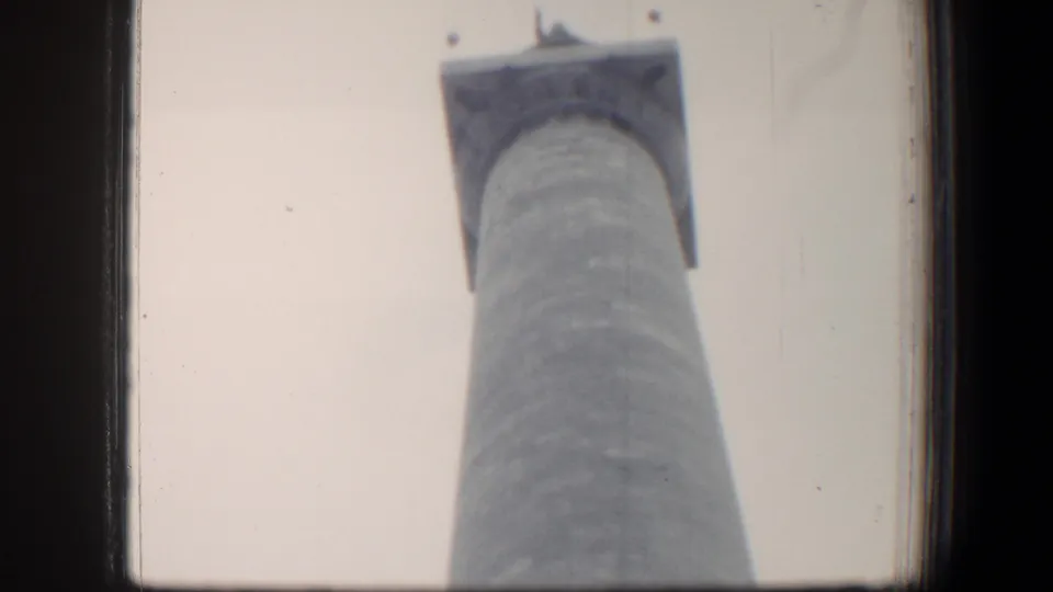 Low-angle 1960 Baltimore home-movie frame of a tall cylindrical monument rising into a pale sky.