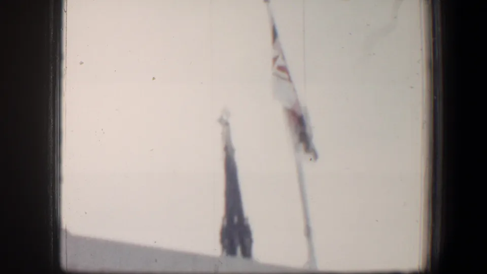 1960 Baltimore 8mm frame with a distant steeple and flag beside a roofline under a pale cloudy sky.