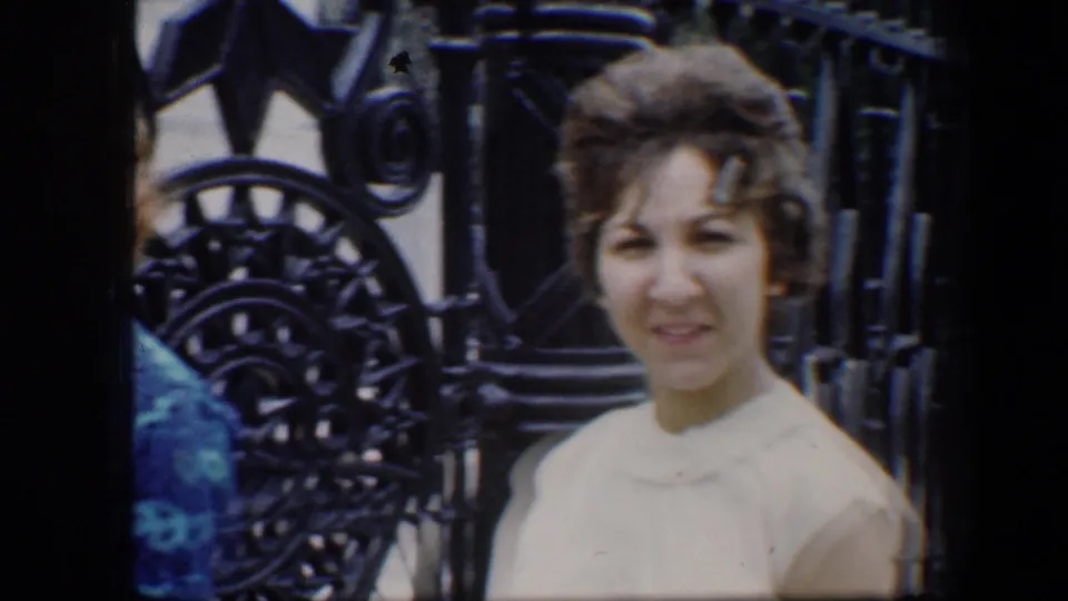 1960 Baltimore home-movie frame of a woman in a light sweater posing near ornate black ironwork at a monument.