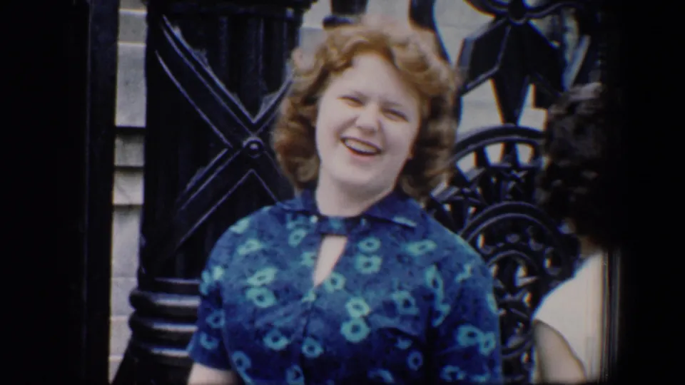 1960 Baltimore home-movie frame showing a smiling woman in a blue patterned dress beside ornate monument fencing.