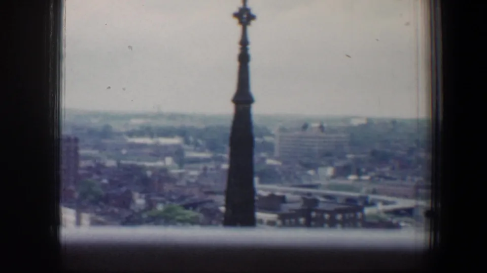 1960 Baltimore skyline seen through a high window, with a dark spire in the foreground and hazy rooftops beyond.