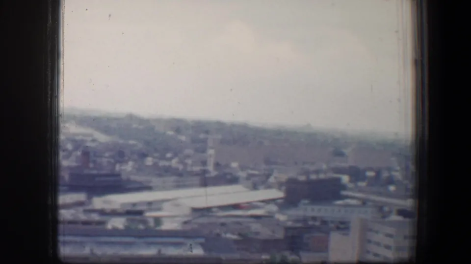1960 Baltimore cityscape viewed through a dark window or railing, with low buildings, rooftops, and a hazy distant skyline.