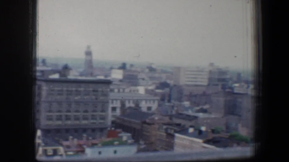 1960 Baltimore cityscape viewed through a high window, with masonry buildings, rooftops, and a hazy skyline beyond.