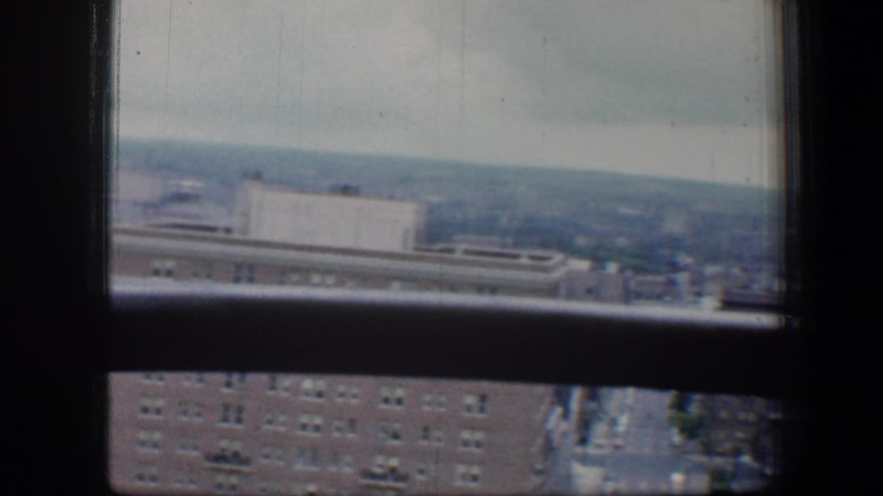 1960 Baltimore city view through a high-rise window, with brick buildings, distant rooftops, and an overcast horizon.