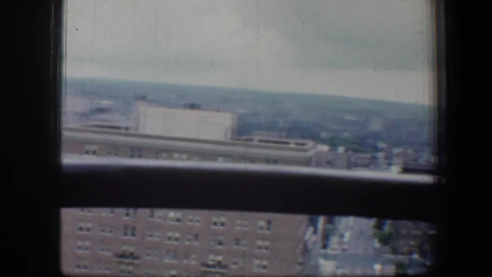 1960 Baltimore city view through a high-rise window, with brick buildings, distant rooftops, and an overcast horizon.