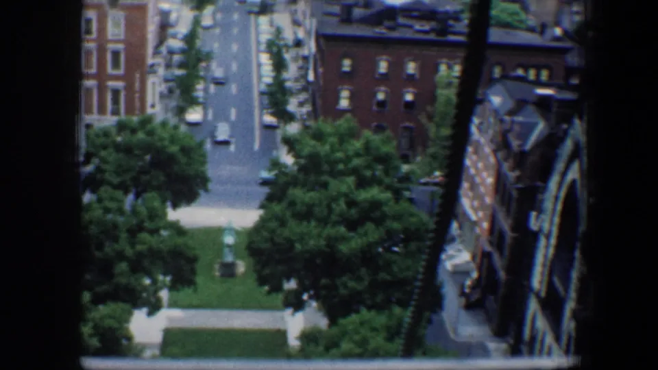 High-angle 1960 Baltimore city view with window mullions, mid-century cars, brick facades, and a small green square.