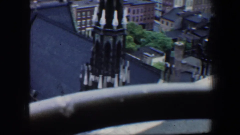 1960 Baltimore home-movie frame looking down past a dark window edge toward a church spire and dense city buildings.