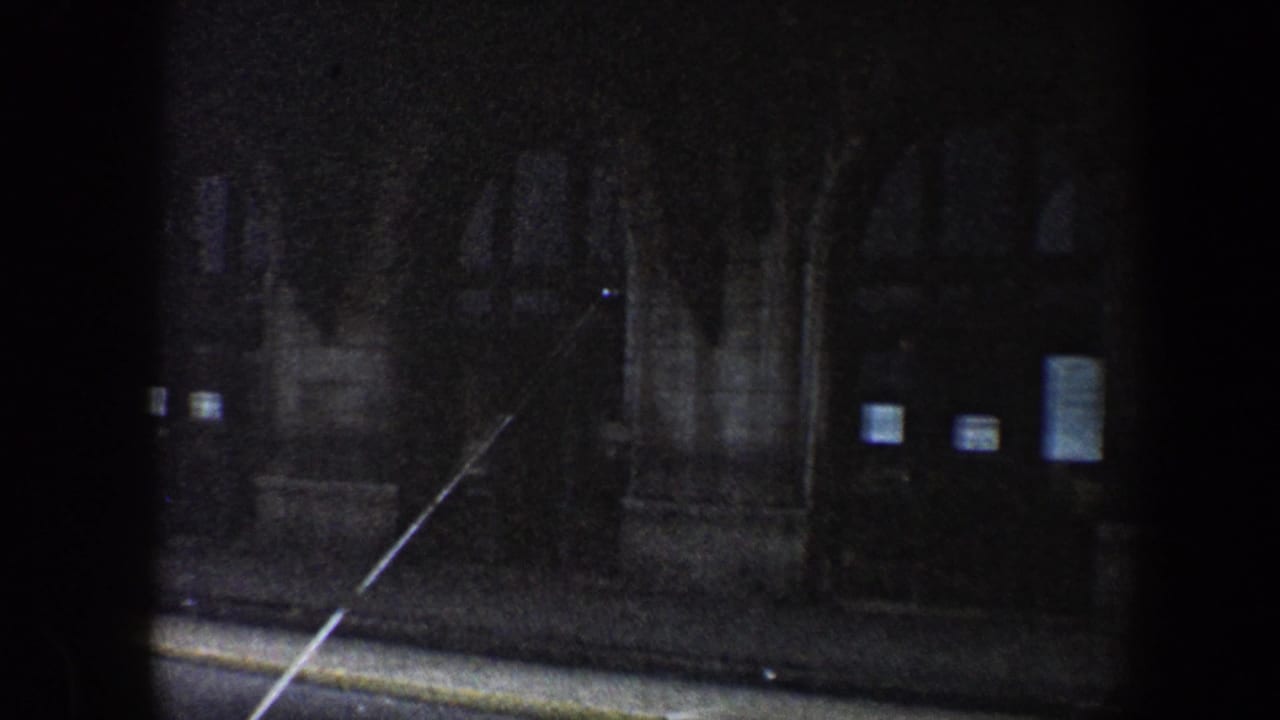1960 Baltimore night street view from a moving vehicle, showing a dark masonry building, curb, and small lit windows.