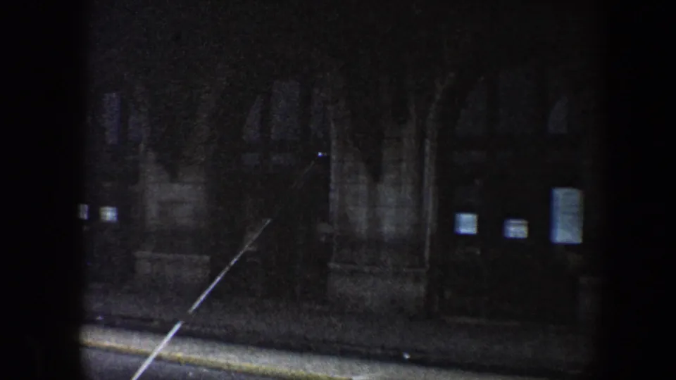 1960 Baltimore night street view from a moving vehicle, showing a dark masonry building, curb, and small lit windows.