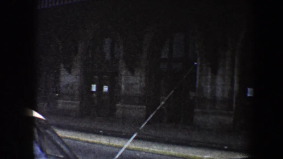 Moving 8mm view of a Baltimore street at night with a dark building facade, curb, and foreground obstruction.