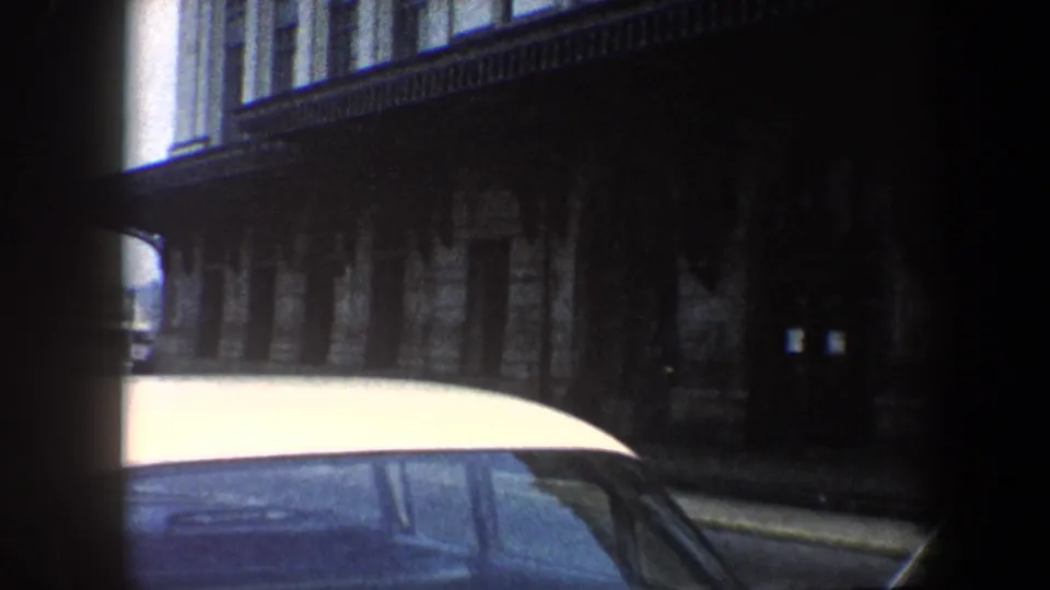 Low-light 8mm frame of a parked white car and dark Baltimore building facade from a moving vehicle.