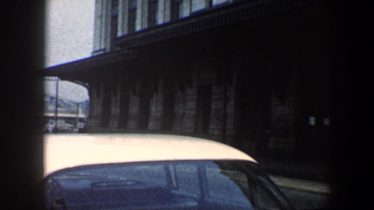 1960 Baltimore 8mm frame showing a white-roofed car passing a shadowed stone building with upper windows.