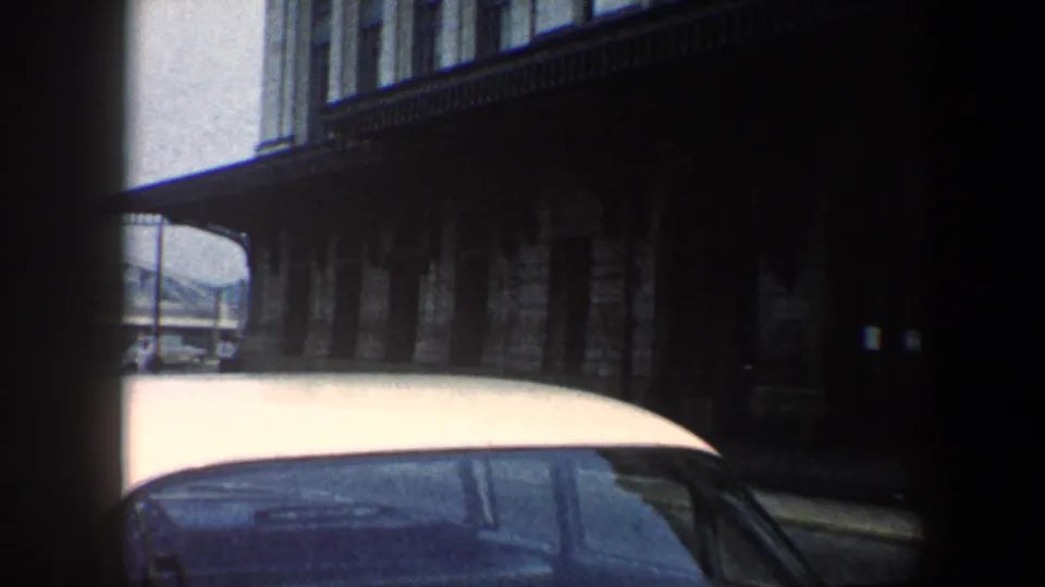 1960 Baltimore 8mm frame showing a white-roofed car passing a shadowed stone building with upper windows.