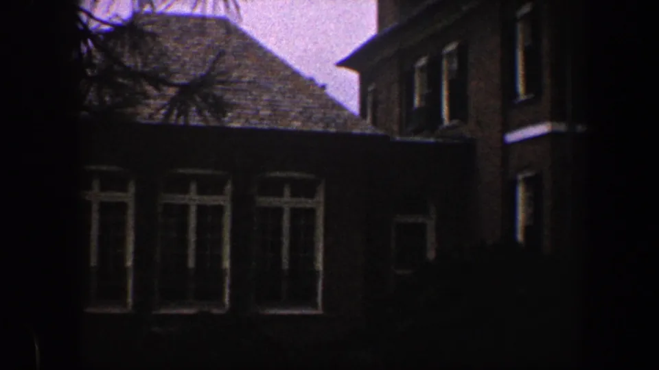 A shadowed brick residential facade with tall white-trimmed windows, seen in grainy 8mm footage from Baltimore metadata.