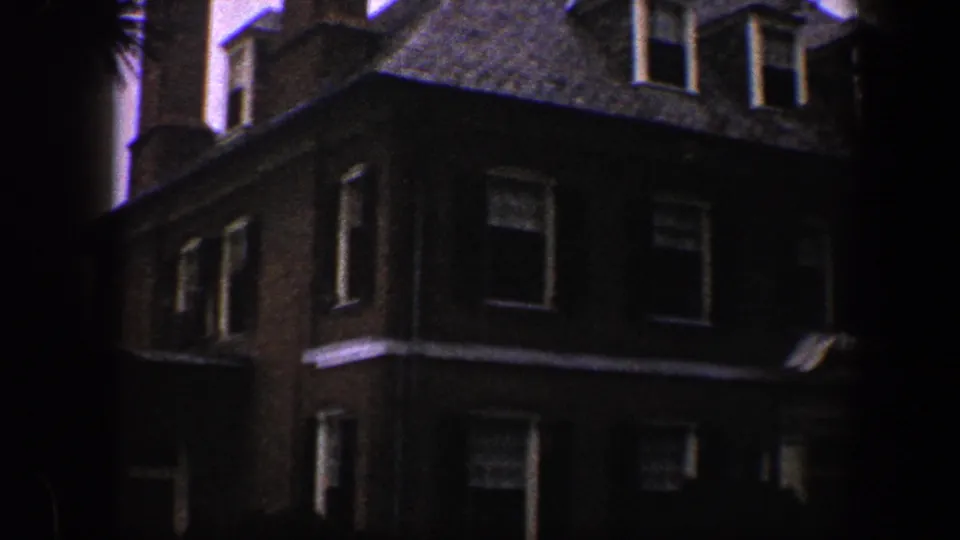 1960 Baltimore 8mm frame showing the upper facade of a dark brick multi-story house with dormer windows and surrounding trees.